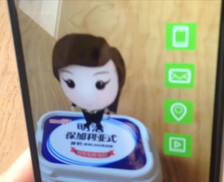 In einem Mobiltelefon ist die Kamera an und zeigt auf eine Joghurtverpackung auf der ein kleiner Avatar steht. Neben dem Augmented Reality Avatar erscheinen Menüpunkte zum clicken.  