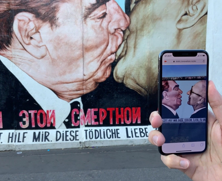 An der Berliner Mauer ist ein Bild von 2 küssenden Männern. Es handelt sich um Leonid Breschnew und Erich Honecker. Vor dem Bild an der Mauer filmt jemand mit einen Handy wie sich die Münder der beiden Männer nähern. Es ist eine Animation in Augmented Reality die in einem Webbrowser zu sehen ist.